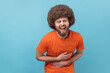 © khosrork - Portrait of laughing man with Afro hairstyle in orange T-shirt holding his stomach and hunched in crazy hysterical laughter, sincere joyful emotions. Indoor studio shot isolated on blue background.