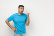 Portrait of amazed shocked young adult unshaven man wearing blue T- shirt standing indicating at advertisement area, copy space for promotion. Indoor studio shot isolated on gray background.