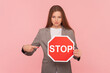 Portrait of serious strict woman with brown hair pointing at red stop sign, looking at camera with bossy expression, forbidden, wearing business suit. Indoor studio shot isolated on pink background.