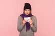 Portrait of sad sick woman with brown hair standing with napkin in hands, suffering flu and running nose, looks unhealthy, wearing hat and scarf. Indoor studio shot isolated on pink background.