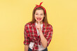 Portrait of positive cheerful woman keeps finger near red lips keeping secret, smiling with cunning look, wearing checkered shirt and headband. Indoor studio shot isolated on yellow background.