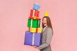 Side view portrait of surprised beautiful woman with brown hair celebrating birthday, holding stack of presents, wearing business suit. Indoor studio shot isolated on pink background.