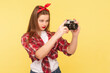 Portrait of concentrated woman photographer with red lips standing with camera in hands, wearing checkered shirt and headband. Indoor studio shot isolated on yellow background.