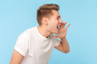 Side view portrait of angry aggressive man wearing white t-shirt standing screaming loud, making announce, arguing with somebody. Indoor studio shot isolated on blue background