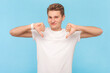 Portrait of frustrated disappointed man wearing white t-shirt looking at camera with frowning face, showing dislike gesture, thumbs down. Indoor studio shot isolated on blue background