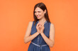 Portrait of sly brunette woman wearing denim dress looking at camera with cunning smile having devious idea preparing prank. Indoor studio shot isolated on orange background.