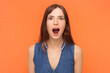 Portrait of amazed shocked surprised brunette woman looking at camera with big eyes and open mouth, sees something astonished, wearing denim dress. Indoor studio shot isolated on orange background.