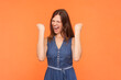 Portrait of overjoyed screaming happy brunette woman wearing denim dress clenched fists celebrating goal achievement rejoicing. Indoor studio shot isolated on orange background.