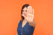 Portrait of serious strict unhappy brunette woman wearing denim dress showing stop sign ban prohibition gesture rejecting. Indoor studio shot isolated on orange background.