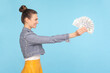 Side view portrait of smiling positive rich woman with bun hairstyle wearing denim jacket giving holding out fan of dollars banknotes. Indoor studio shot isolated on light blue background.