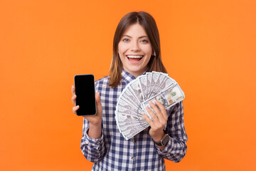 Portrait of smiling happy positive woman with brown hair showing smartphone with empty display and dollar banknotes, wearing checkered shirt. Indoor studio shot isolated on orange background