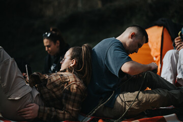 Wall Mural - A group of young adults relaxes at a campsite, with one person checking a phone, highlighting a connection amidst a natural setting.