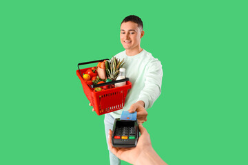 Poster - Young man with full shopping basket paying via terminal on green background