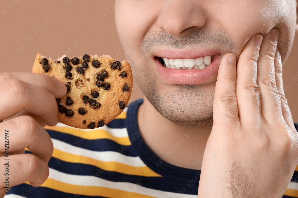 Young man with cookie suffering from toothache on brown background, closeup