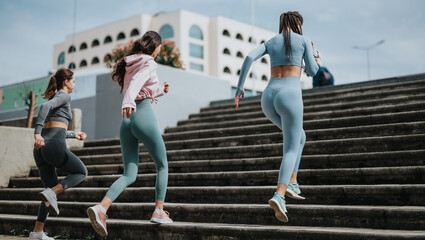 Wall Mural - Active young women in workout attire exercising together by running up city stairs, showcasing health and companionship.