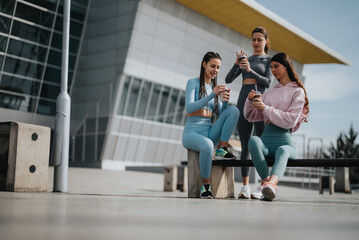 Wall Mural - Three athletic women in sportswear relaxing and checking phones after a workout session outside a contemporary fitness center.