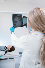 female doctor examines pictures of the skull in the x-ray room, in the background lies a patient with a traumatic brain injury and concussion