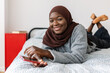 Portrait of beautiful young muslim black woman in brown headscarf holding brand smart phone device while smiling at camera lying on bed.