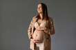 Smiling dreamy brown haired young pregnant woman wearing beige jacket, bra, leggings holding hands on her bare belly looking away with satisfied facial expression