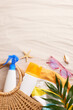 A top-view vertical photo of beach vacation items, including sunscreen bottles, sunglasses and a straw bag, arranged on a sandy backdrop