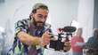 Young Latin Male Filmmaker With Beard And Glasses, Joyfully Operating A Camera In A Busy Studio, Surrounded By Diverse Team Members Focused On A Creative Video Production Project.