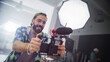 © Gorodenkoff - Young Caucasian Male Videographer With Beard, Wearing Colorful Shirt And Cap, Enthusiastically Operates Camera In Studio Setting, Capturing Content For Advertising Or Movie Production.