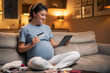 A joyful pregnant woman using a tablet to shop online for baby goods while sitting on the couch in the living room