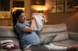 A happy pregnant woman having a video call on her laptop with her family and showing the baby clothes she had bought