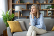 © Liubomir - A thoughtful senior woman sits alone on a couch, looking away with a reflective expression in a well-furnished living room surrounded by books and plants.