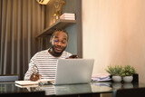 Fototapeta Nowy Jork - Smiling male entrepreneur working with laptop and making note at desk in living room