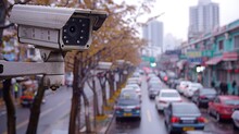 A Surveillance Camera Is Mounted On A Building, Overlooking A Busy Street With Cars And People Passing By.
