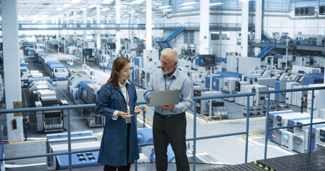 Aufkleber - Two Multiethnic Engineers Standing on a Platform, Using Laptop Computer at an Electronics Factory. Machines are Undergoing Maintenance, Specialists Monitoring the Progress Through Online Software