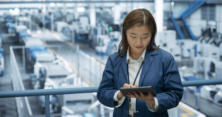 Fototapete - Young Asian Female Engineer Standing on a Platform, Using Tablet Computer at an Electronics Factory. Machines are Undergoing Maintenance, Specialist Monitoring the Progress Through Online Software
