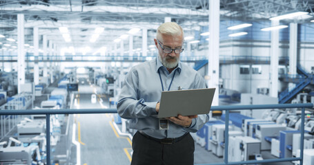 Fototapete - Senior Engineer Standing on a Platform, Using Laptop Computer at an Electronics Factory. Machines are Undergoing Maintenance, Specialist Monitoring the Updates Through Online Software