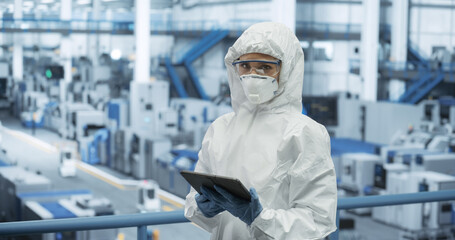 Leinwandbilder - Female Worker Wearing Full-Body Protective Coveralls, Mask and Goggles at a Manufacturing Plant. Portrait of a Young Woman Specialists Using a Tablet Computer, Looking at Camera, Smiling
