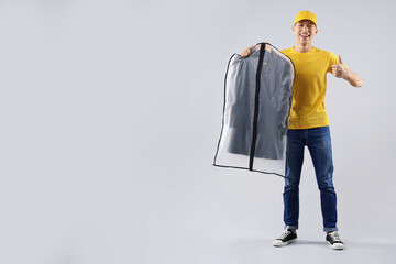 Wall Mural - Dry-cleaning delivery. Happy courier holding garment cover with clothes and showing thumbs up on light grey background, space for text