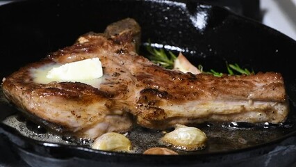 Wall Mural - A juicy pork cutlet on the bone with a piece of butter, garlic and rosemary is fried in a frying pan. Close-up. Super slow motion.