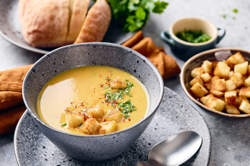 Wall Mural - Bowl of mashed potato soup with a spoon on the plate. The bowl is filled with yellow soup puree and topped with croutons, parsley and ground red pepper.