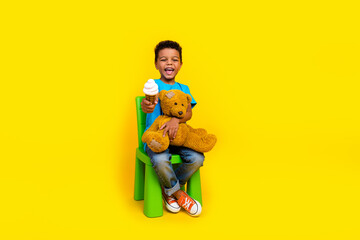 Wall Mural - Full size photo of positive cheerful cute schoolboy wear blue t-shirt hold teddy bear give you ice cream isolated on yellow background