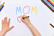 Kids girl draws the word mom on a paper sheet with markers and drawing a postcard for mom, top view.