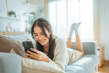 Fototapeta Mapy - Happy young asian woman lady browsing surfing wireless internet on mobile phone while sitting a sofa couch in living room at home apartment, Shopping online via website