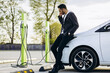 Man at charging station charging electric car and using mobile phone
