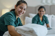 © AI_images - A cheerful Latina maid and her female co-worker are seen making the bed in a hotel room, exuding positivity and teamwork.