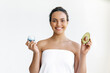 Skincare concept. Beautiful charming well-groomed brazilian or hispanic young woman wrapped in a towel stands on white background, holds an avocado and moisturizing cream in her hands, smile at camera