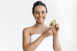 Charming well-groomed brazilian or hispanic young woman wrapped in a towel stands on a white background, holds an avocado in her hands, looks and smiles at the camera
