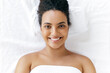 Top view of a beautiful well-groomed brazilian or hispanic young woman wrapped in a towel, lying in a beauty salon before a massage or facial skin care procedure, looking at the camera, smiling