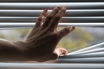 Man separating slats of white blinds indoors, closeup