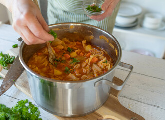 Wall Mural - Cooking pot with fresh cooked healthy vegetables soup. Sprinkling chopped parsley over the soup