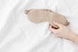 Beige eye mask for sleep on bed background, minimal lifestyle aesthetic, copy space. Top view woman hand holding sleeping mask on white bed linen, morning daylight. Comfort rest, healthy trend