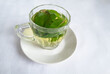 Mint tea leaves in hot water in a glass mug with a handle on a white saucer. The table has a white paper tablecloth.